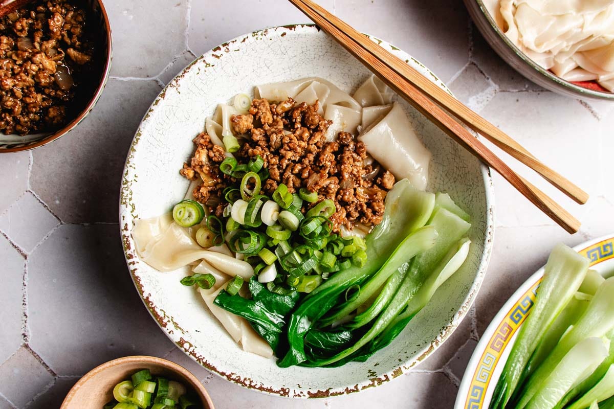 A bowl of wonton skin noodles topped with minced meat, bok choy, and chopped green onions is served as a delightful wonton wrapper noodle bowl. Chopsticks rest on the bowl, while dishes with extra toppings and bowls are nearby on a tiled surface.