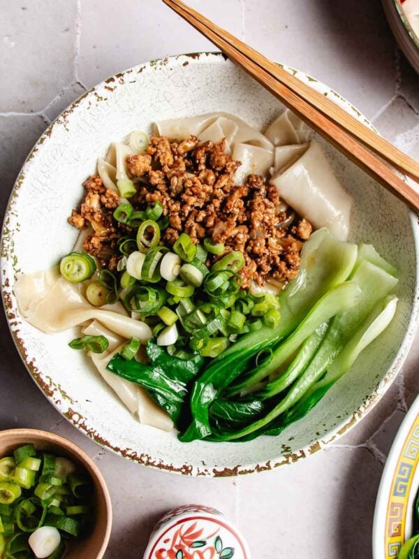 A Wonton wrapper noodle bowl featuring wide noodles topped with ground meat, sliced green onions, and bok choy, with chopsticks resting on the bowl.