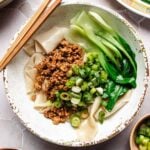 A bowl of wide noodles, made from wonton wrapper noodle bowl, topped with minced meat, sliced green onions, and bok choy, with chopsticks resting on the bowl’s edge.