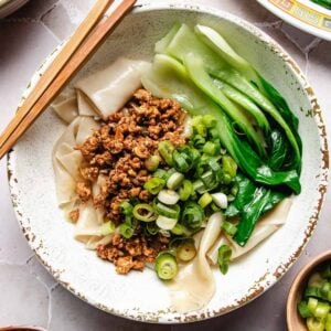A bowl of wide noodles, made from wonton wrapper noodle bowl, topped with minced meat, sliced green onions, and bok choy, with chopsticks resting on the bowl’s edge.