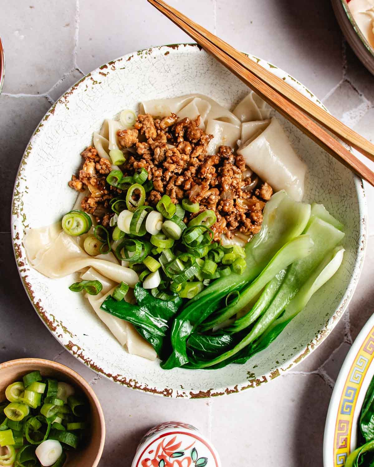 A Wonton wrapper noodle bowl featuring wide noodles topped with ground meat, sliced green onions, and bok choy, with chopsticks resting on the bowl.