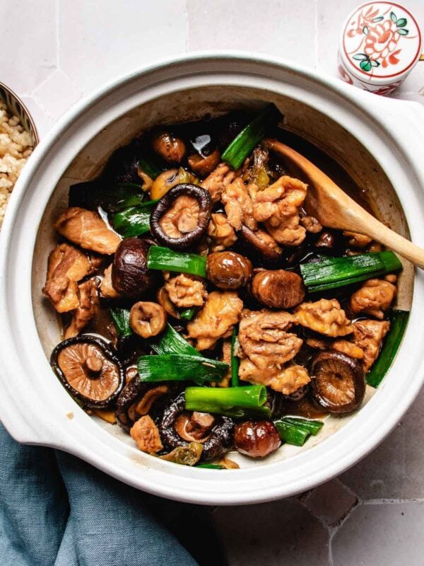 A white ceramic pot filled with Chinese braised chicken with chestnuts, shiitake mushrooms, and green onions, served with a wooden spoon.