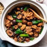 A white clay pot containing Chinese braised chicken with chestnuts, braised with whole shiitake mushrooms and green onions, with a wooden spoon resting inside.