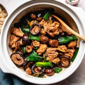 A white clay pot containing Chinese braised chicken with chestnuts, braised with whole shiitake mushrooms and green onions, with a wooden spoon resting inside.