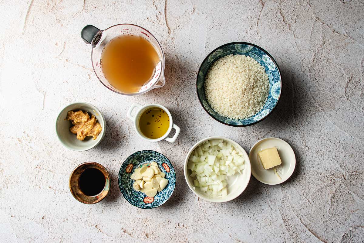 Ingredients for miso mushroom crispy rice including a measuring cup of broth, a bowl of rice, chopped onions, sliced garlic, miso paste, soy sauce, olive oil, and a pat of butter.