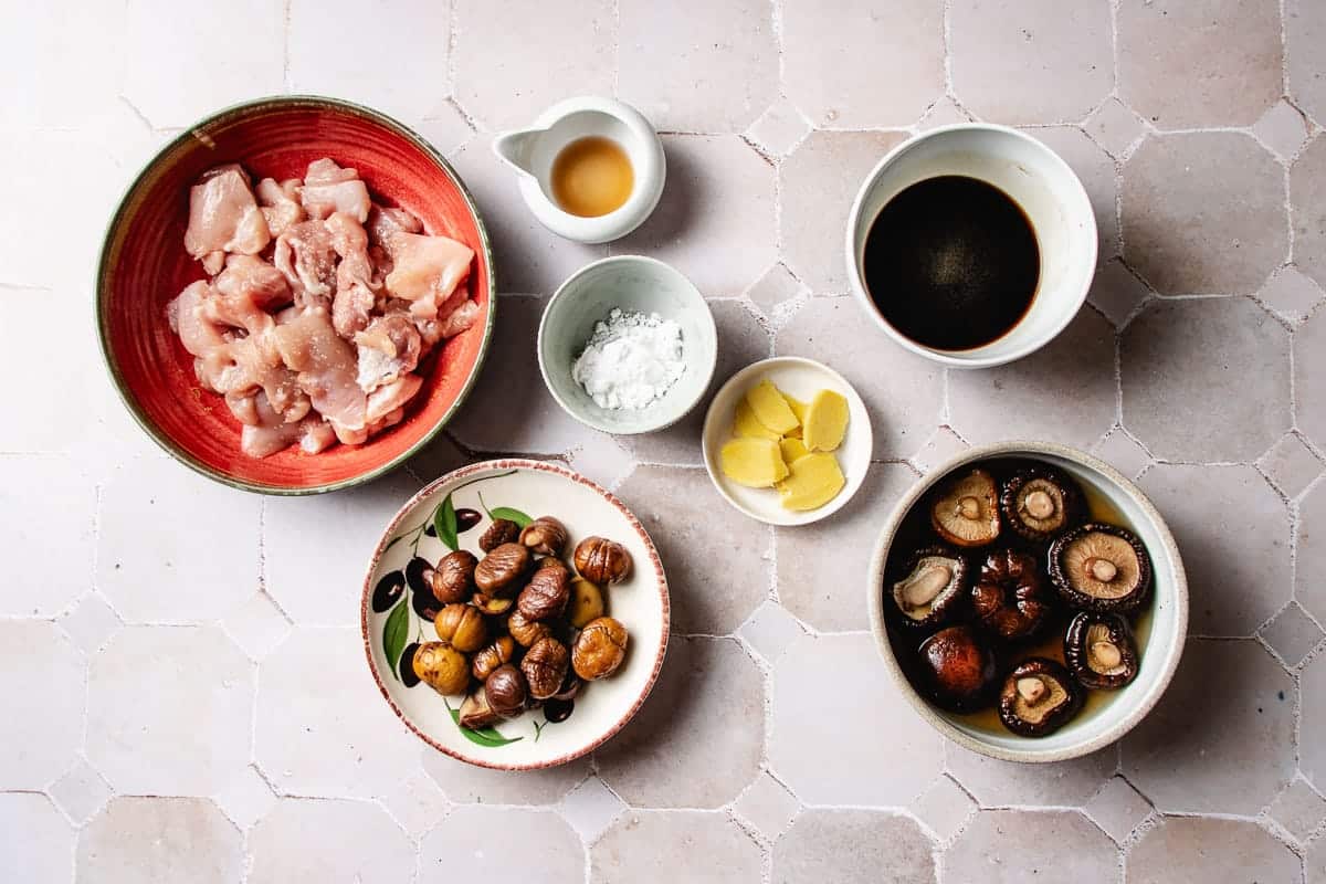 Bowls containing raw chicken pieces, chestnuts, rehydrated shiitake mushrooms, ginger slices, starch, and sauces for Chinese braised chicken with chestnuts, arranged on a tiled surface.
