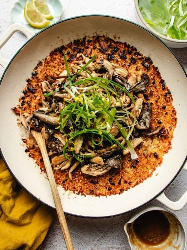 A large skillet of miso mushroom crispy rice topped with sautéed mushrooms and sliced scallions, served with side bowls of lemon wedges and sliced green onions.