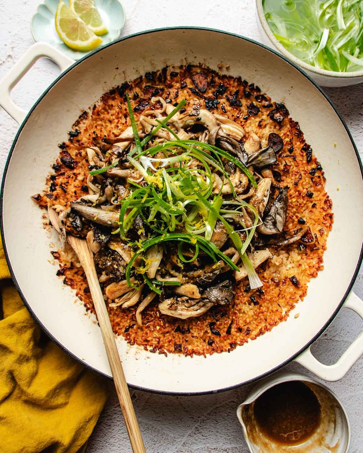 A large skillet of miso mushroom crispy rice topped with sautéed mushrooms and sliced scallions, served with side bowls of lemon wedges and sliced green onions.