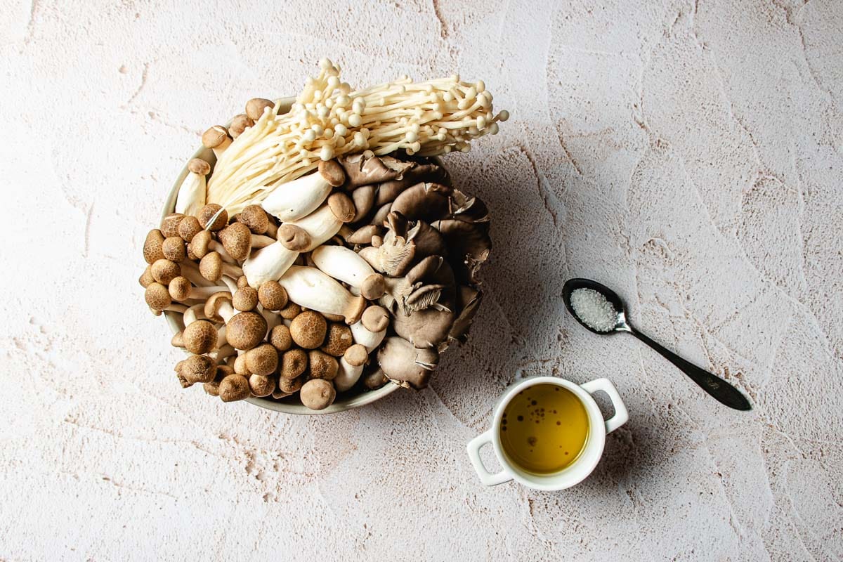 A bowl filled with a variety of raw mushrooms for miso mushroom crispy rice, next to a small bowl of oil and a spoonful of salt.